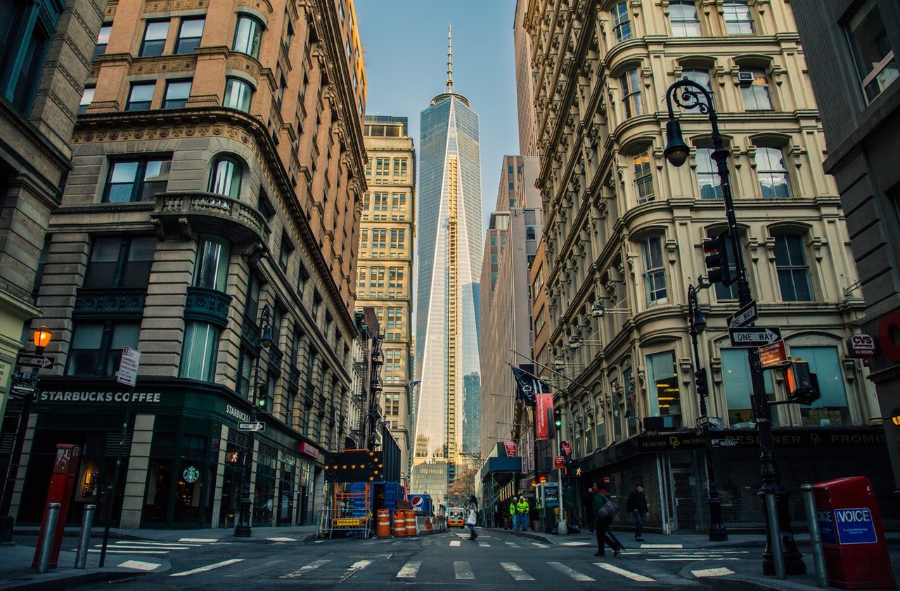 A street scene showcasing tall buildings that frame a view of One World Trade Center in New York City. The foreground features a Starbucks and some construction barriers, while pedestrians stroll along the crosswalk under a clear blue sky.